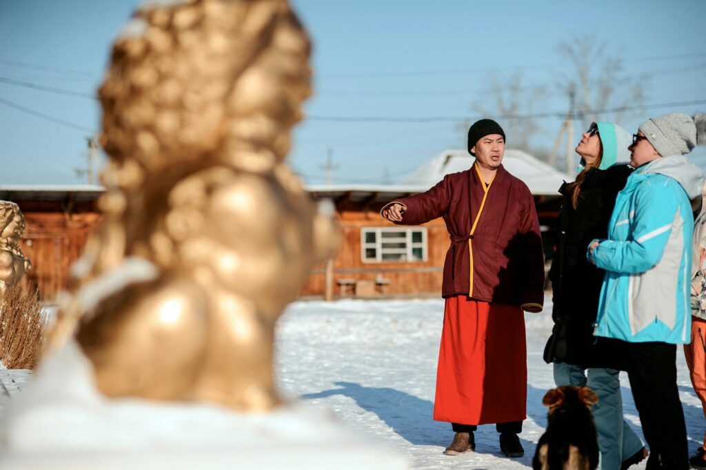 A monk in traditional attire interacts with tourists in a snowy village, with golden sculptures visible.