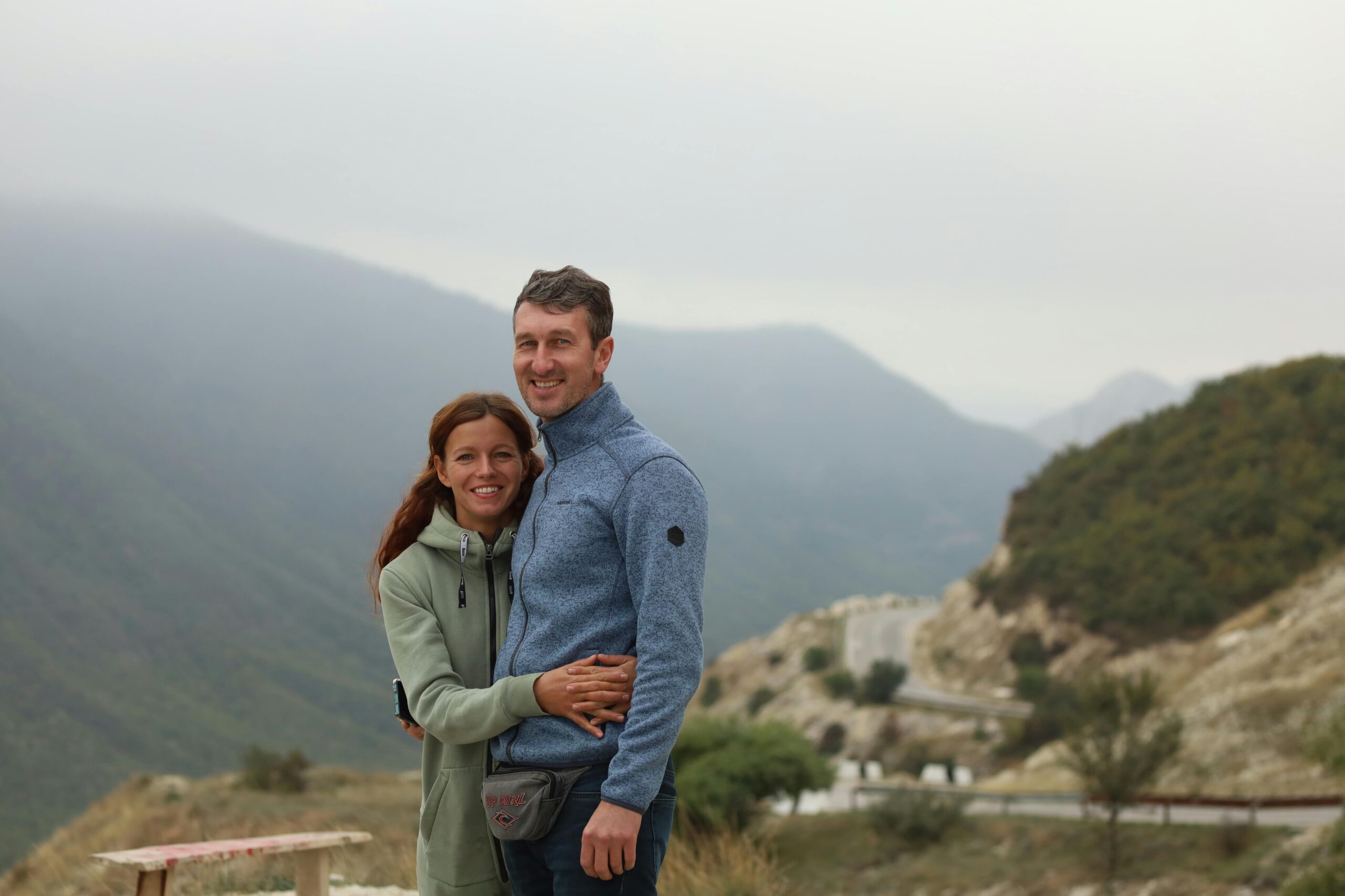 A smiling couple embraces against a stunning mountain backdrop, sharing a joyful travel moment.