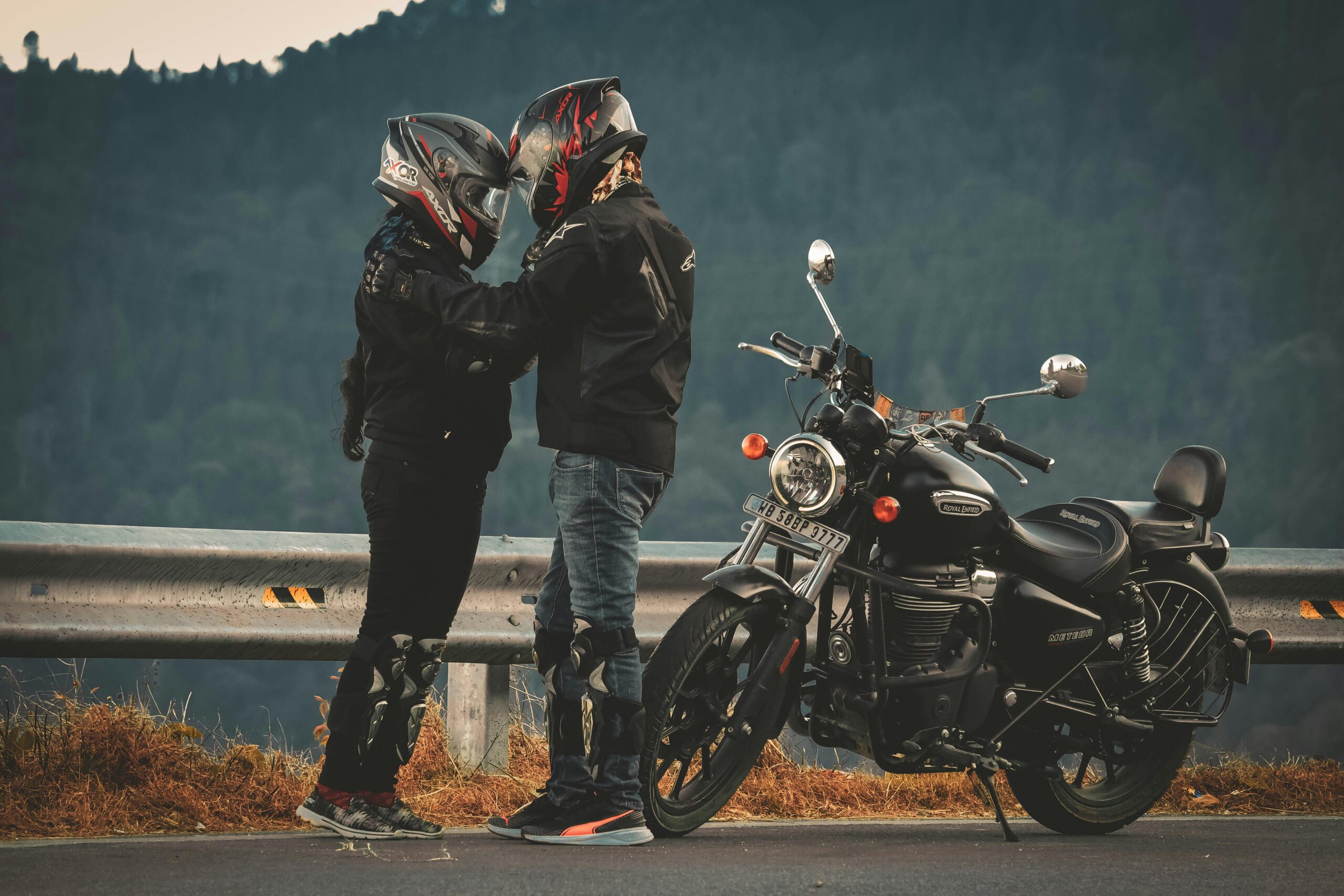 Two motorcyclists in helmets standing by their bike on a scenic road in SK, India.