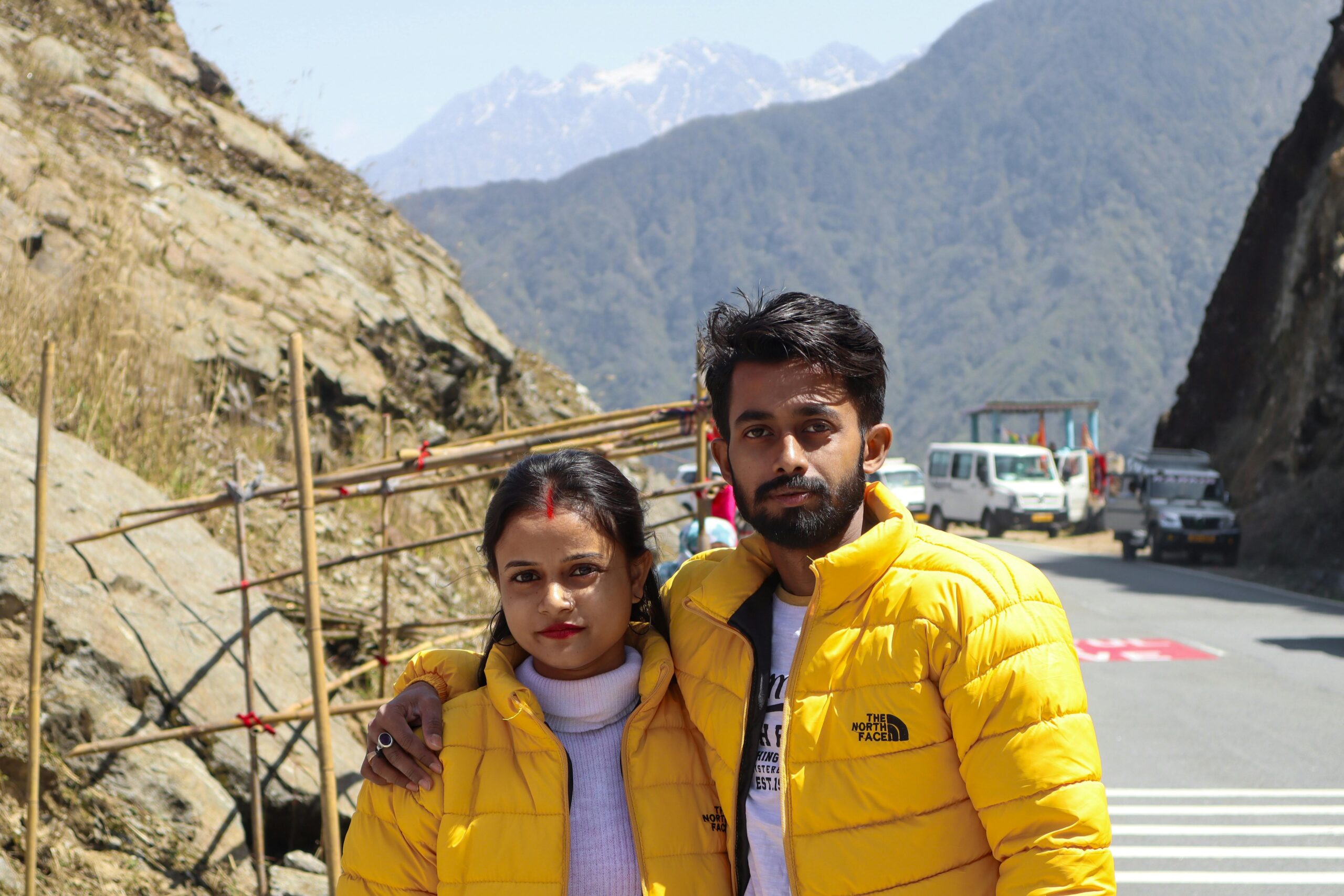 Couple wearing yellow jackets posing on a mountainous road with cars and rocky terrain.
