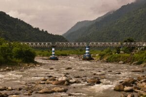 A picturesque view of a river flowing under a steel bridge in India, surrounded by lush mountains.