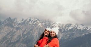 Two women in red shirts and white beanies embracing and smiling against Auli mountains.