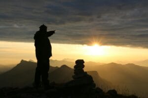 A hiker silhouette pointing during a mountain sunrise with a rock cairn and cloudscape.